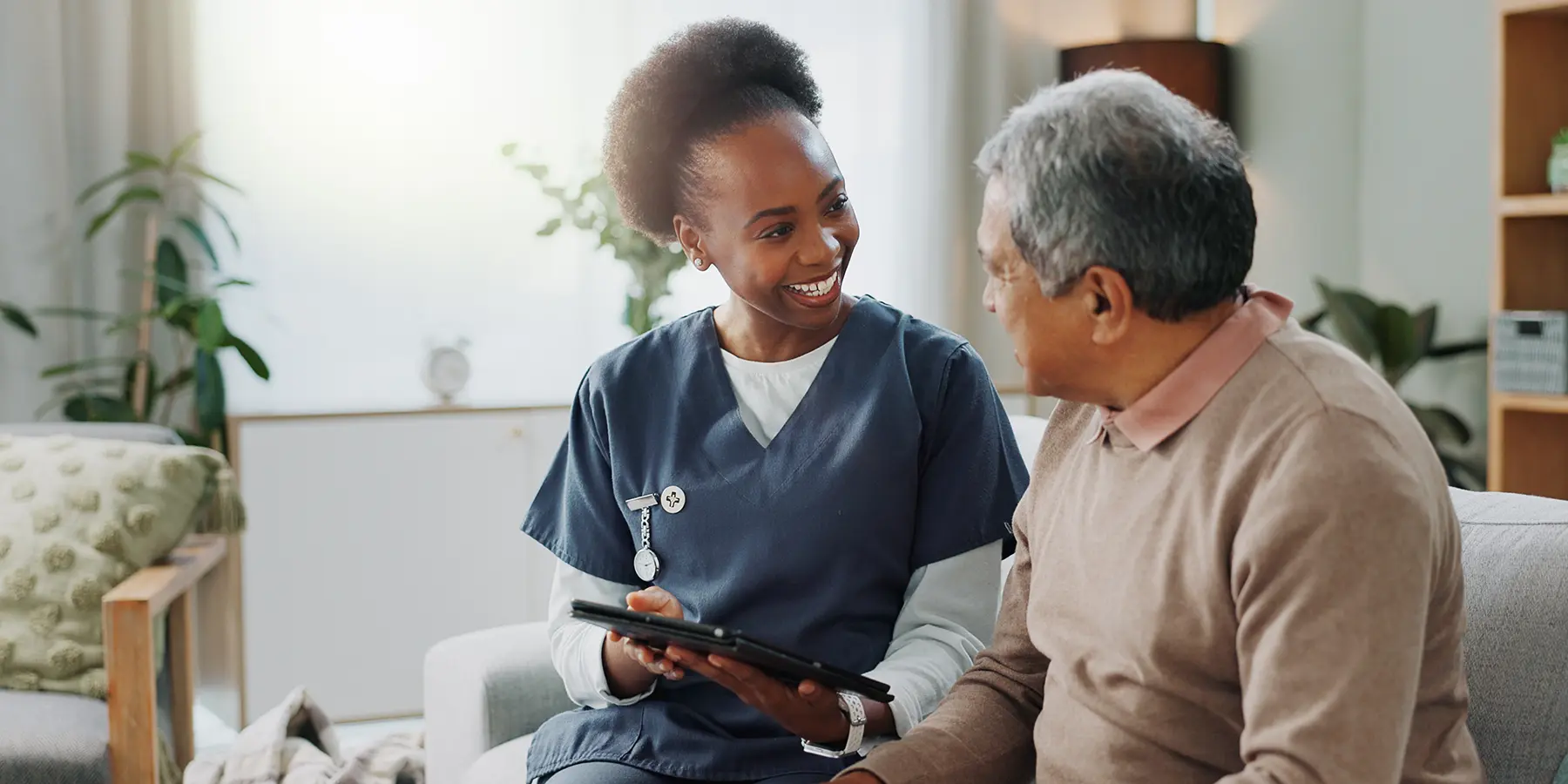 A black nurse with her hair tied back, wearing dark blue scrubs, sits on a sofa and talks to an older man with short dark grey hair who is facing away from the camera