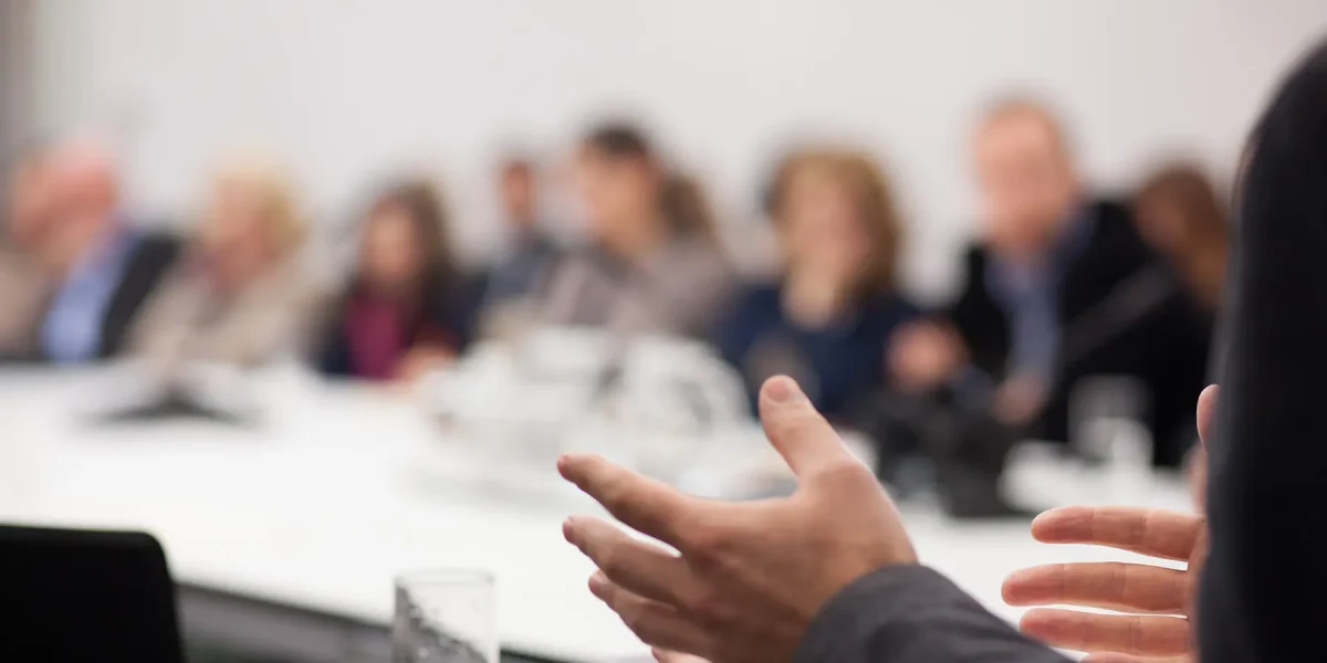 A blurry photo of a meeting, the hands of the person nearest the camera are in focus as they explain something