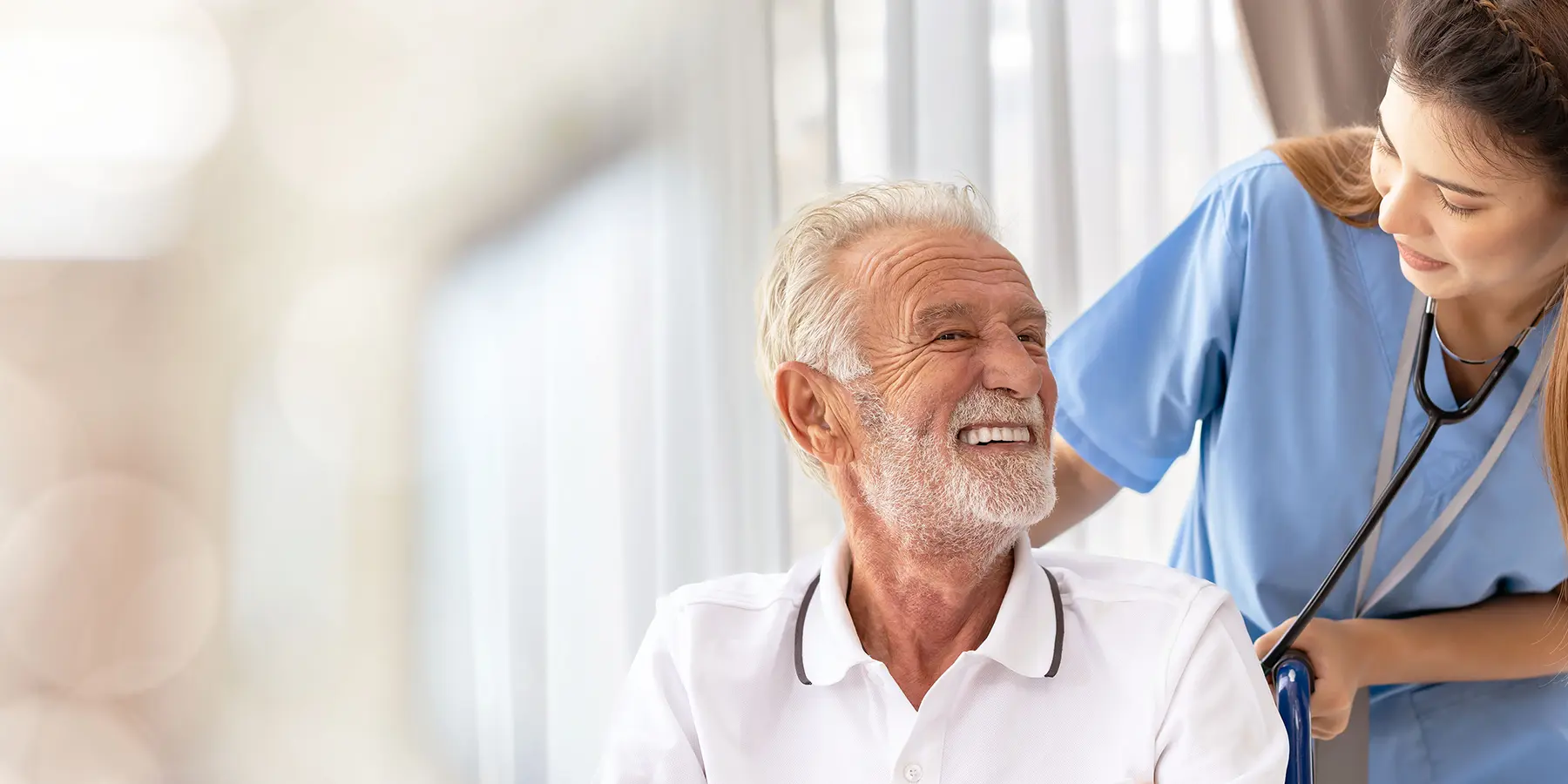 An older white man with white short hair and a beard sits in a chair, and a younger white nurse with long brown hair wearing scrubs leans towards him from behind