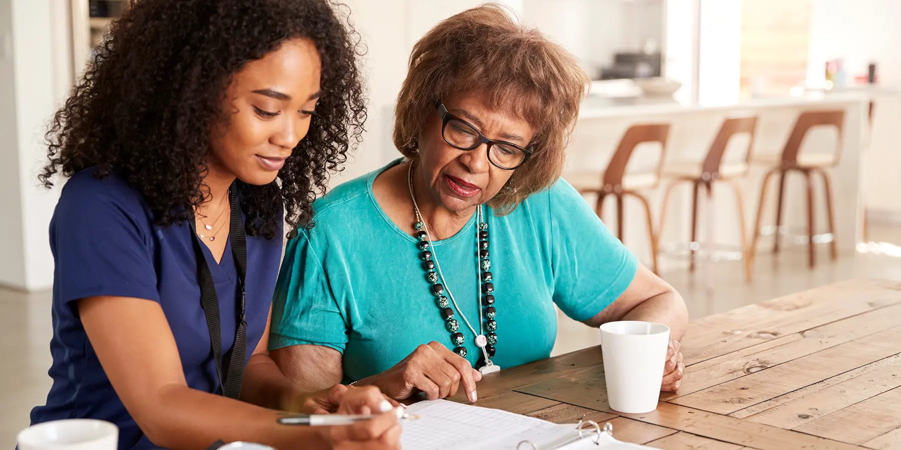 A black nurse with curly hair sits at a table, going over some paperwork with an older black woman with short hair and glasses.