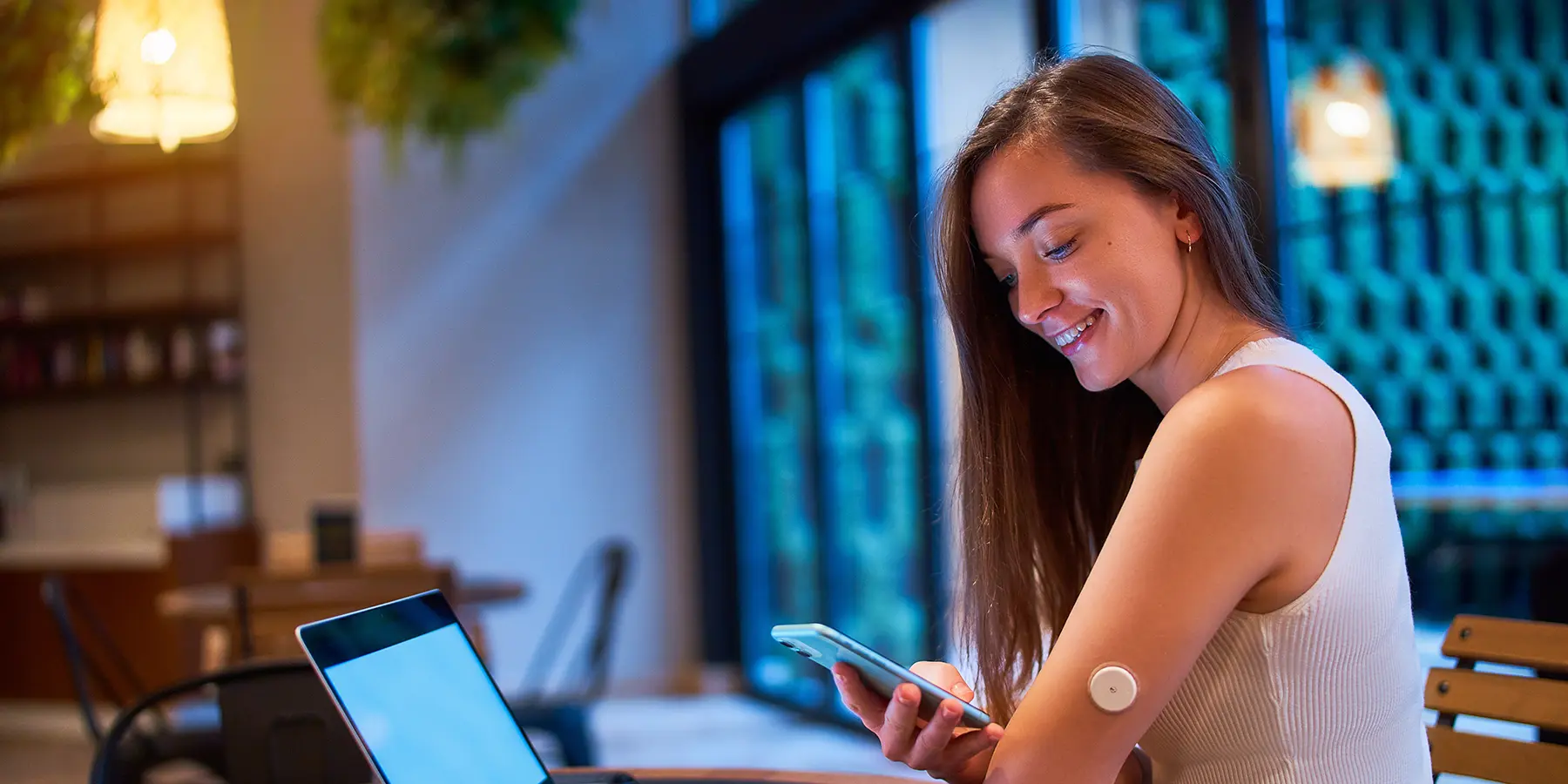 A young white woman wearing a sleeveless white top with long brown hair looks at her phone. On her upper arm is a diabetes monitoring device