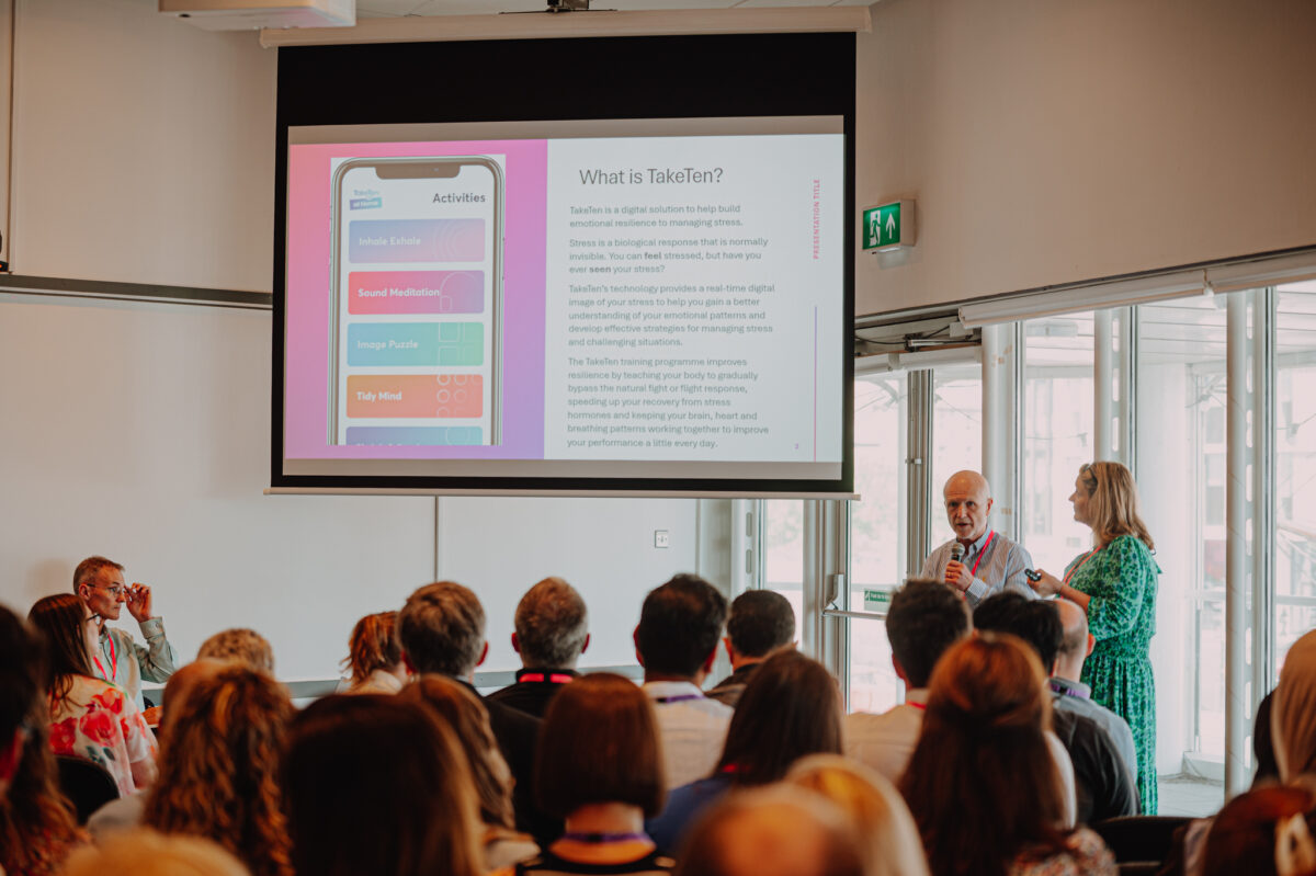 A white man and white woman present at a conference. The man wears a pale shirt, the woman wears a green and white patterned dress. The slide presented shows an overview of the Take Ten app.
