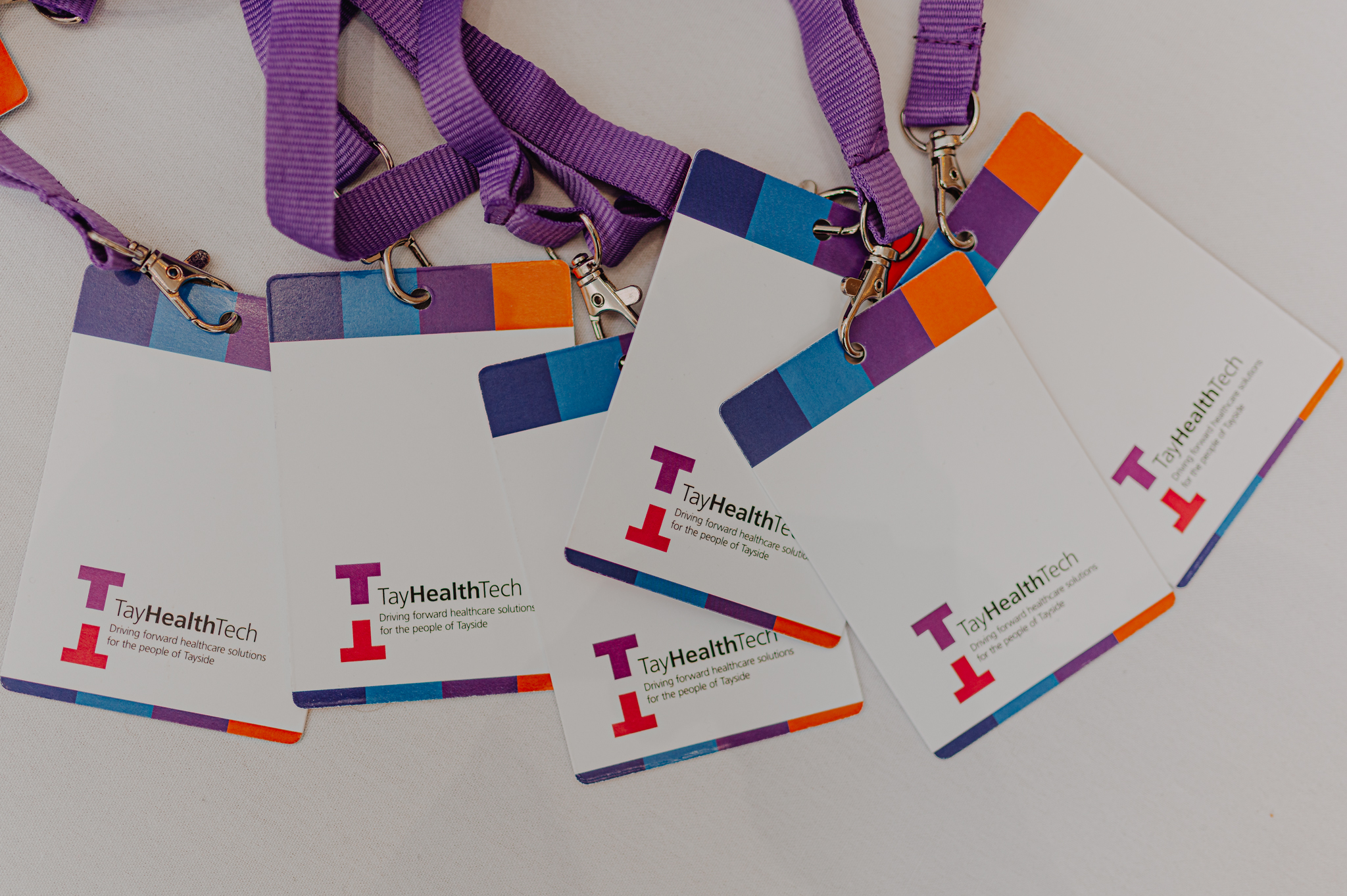 A collection of conference lanyard badges. The lanyards are purple. The badges are white with 4 colour blocks along the top (dark blue, cyan, purple and orange), with the Tay Health Tech logo at the bottom.
