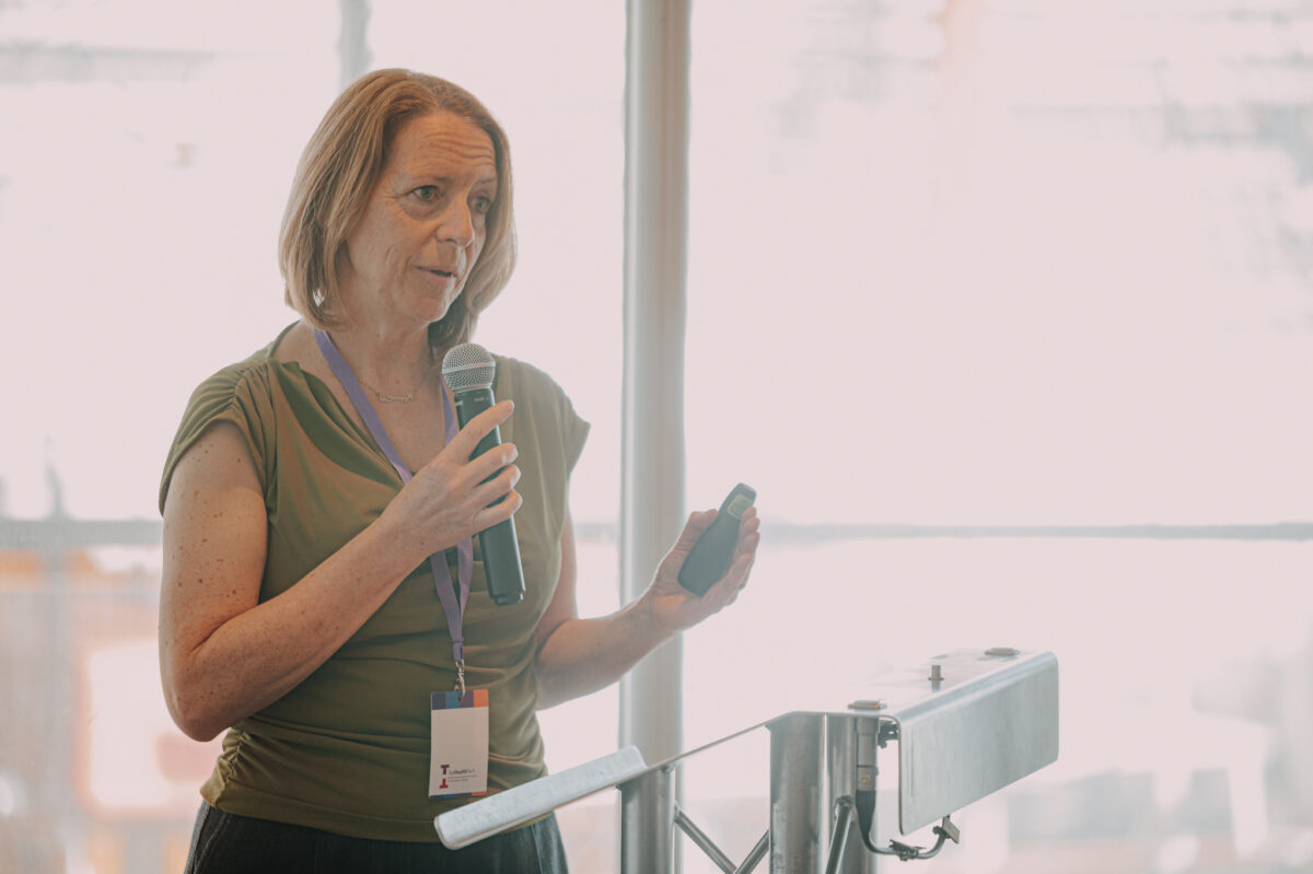 A white woman with medium length dark blonde hair presents from behind the podium. She wears a khaki green cowl neck sleeveless top.