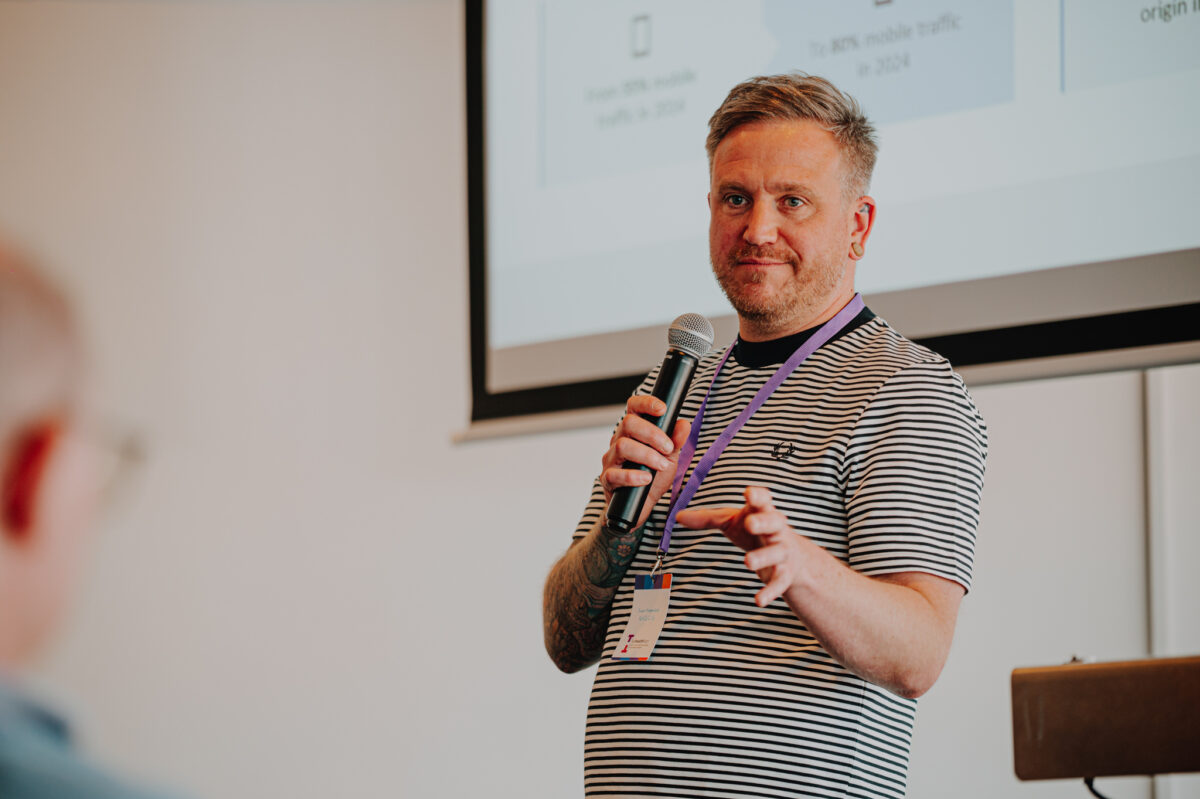 A white man with short blond hair presents in front of the projection screen. He wears a black and white horizontal striped tee.