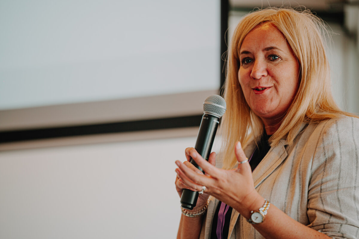 A white woman with long blond hair speaks to an audience, holding a microphone in her right hand. She wears a cream suit.