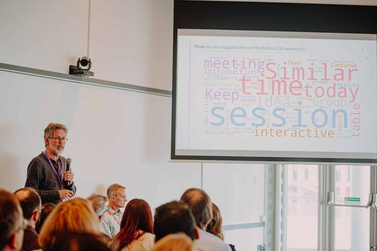 A white man with dark grey hair talks from the front of the conference room, to the side of the projector screen showing a slide about what the Citizens Assembly said was wanted