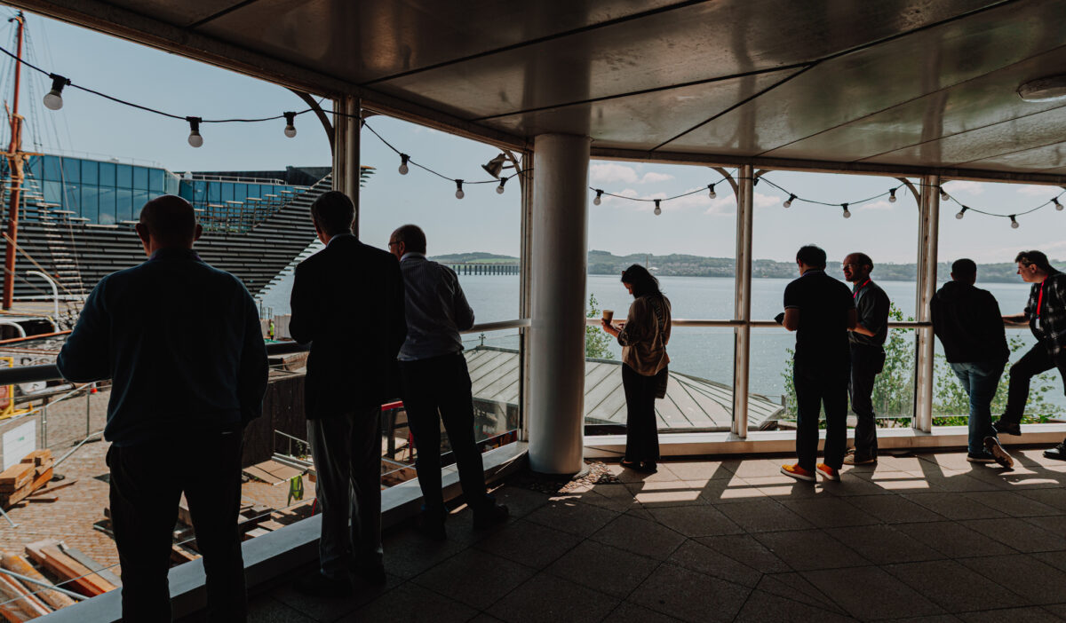 Conference participants stand on a balcony overlooking the River Tay and the V&A Museum. The sun is shining so each person appears as a silhouette.