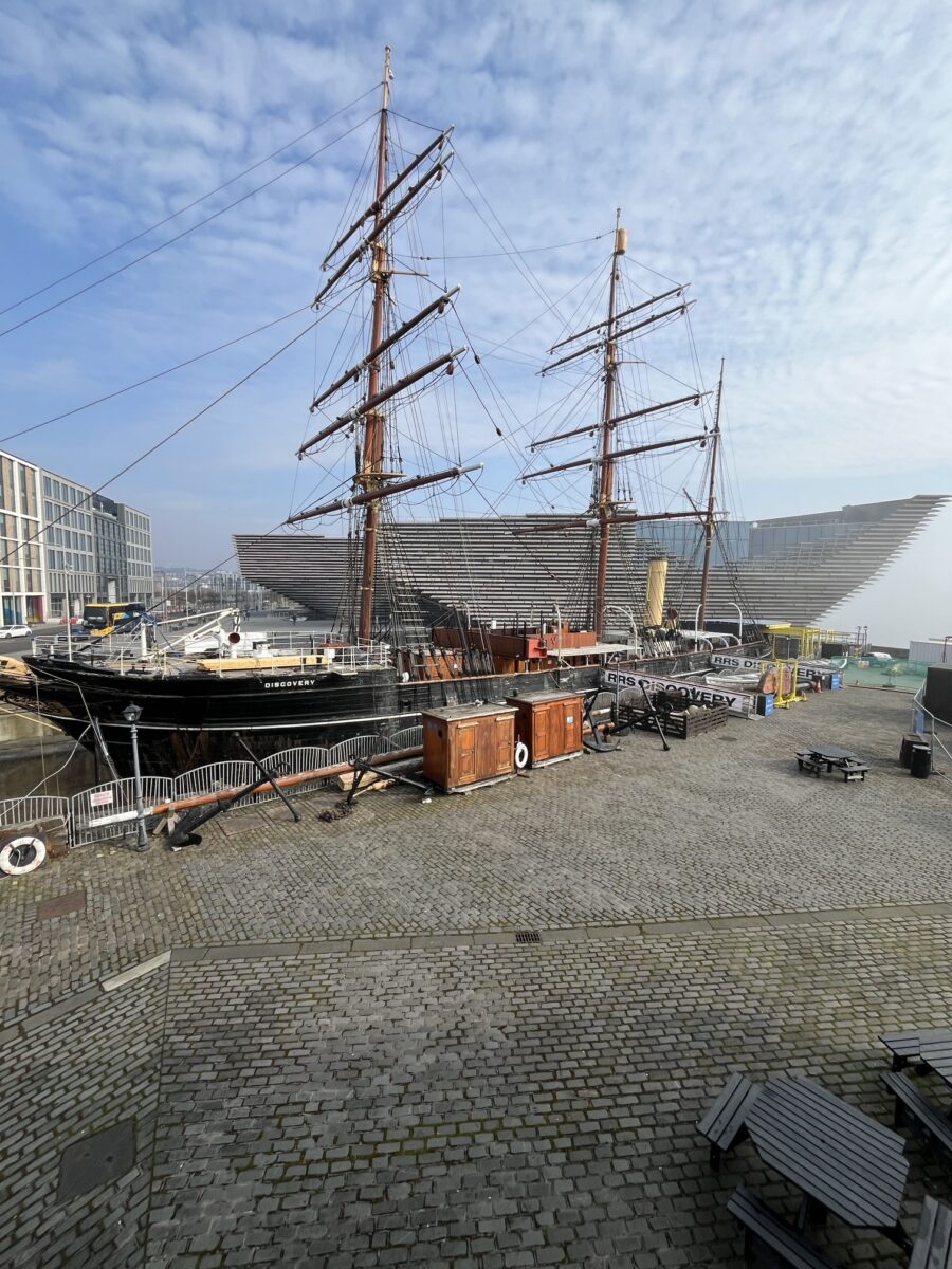 The RRS Discovery at the riverside in Dundee. The ship is a barque-rigged steamship launched in 1901. Behind her is the V&A Dundee museum, under a blue sky.