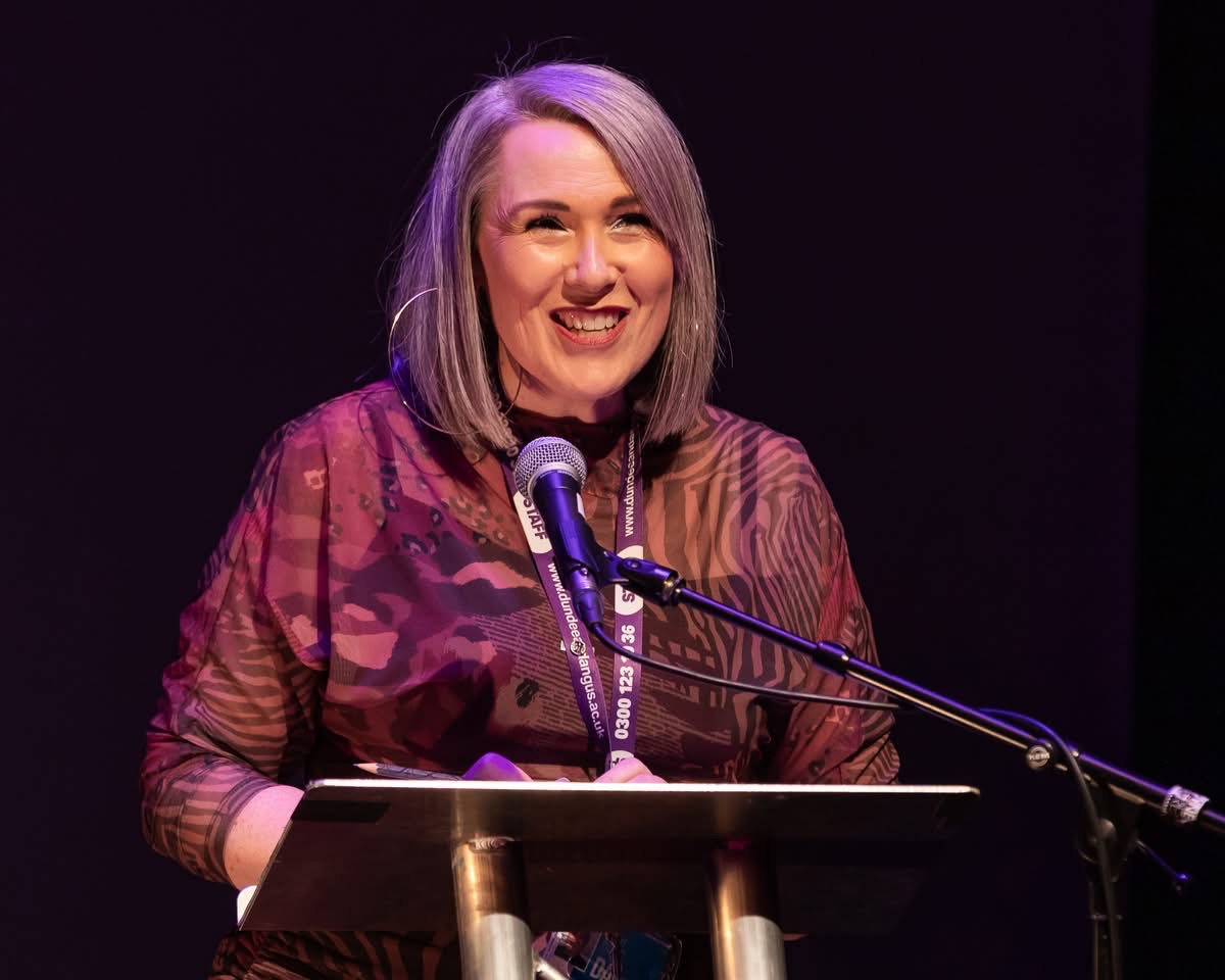 Against a black backdrop, a white woman with mid length silver hair stands behind a lectern and microphone, smiling at an audience behind the camera. She wears large hoop earrings, and an animal print maroon top.