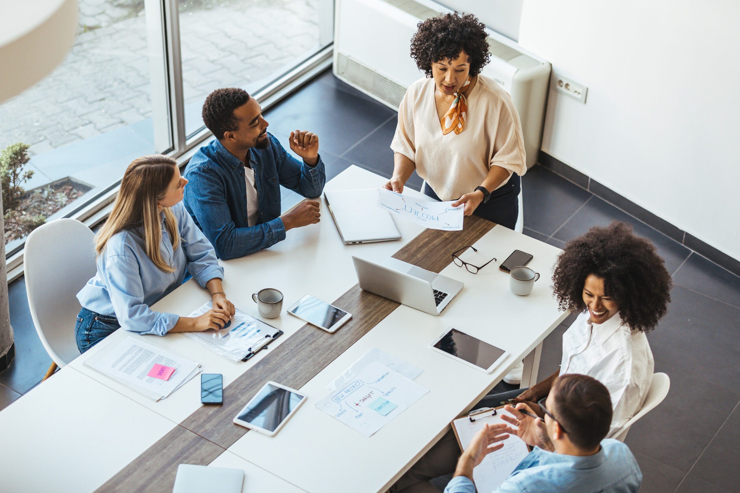 A team captured from above, sit around a large white marble table, each with an electronic device and papers that mention project strategy.