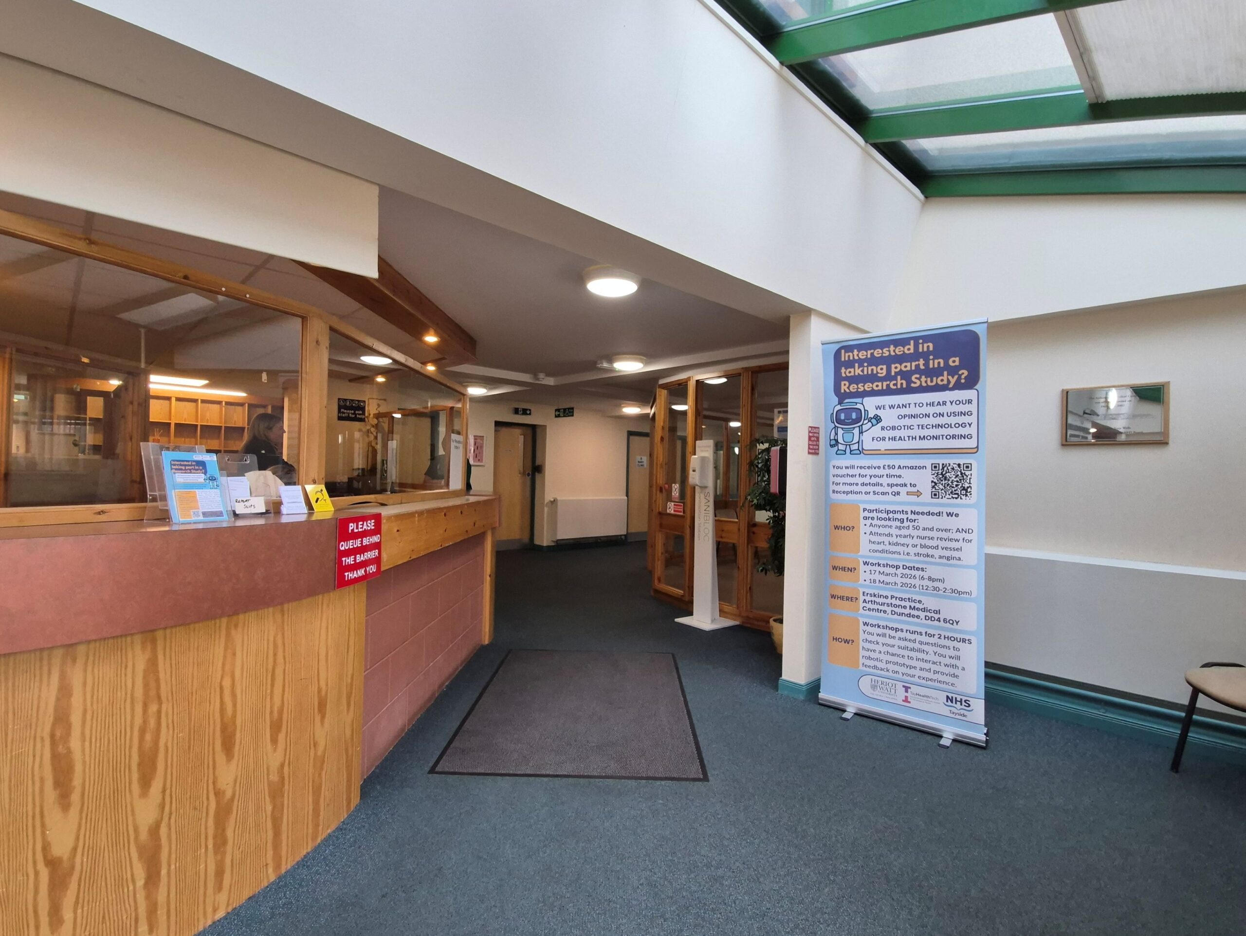 A photo of Erskine GP practice reception area, including the desk on the left, a corridor going back from the centre of the image, and a pop-up banner in the area to the right, promoting a research study.