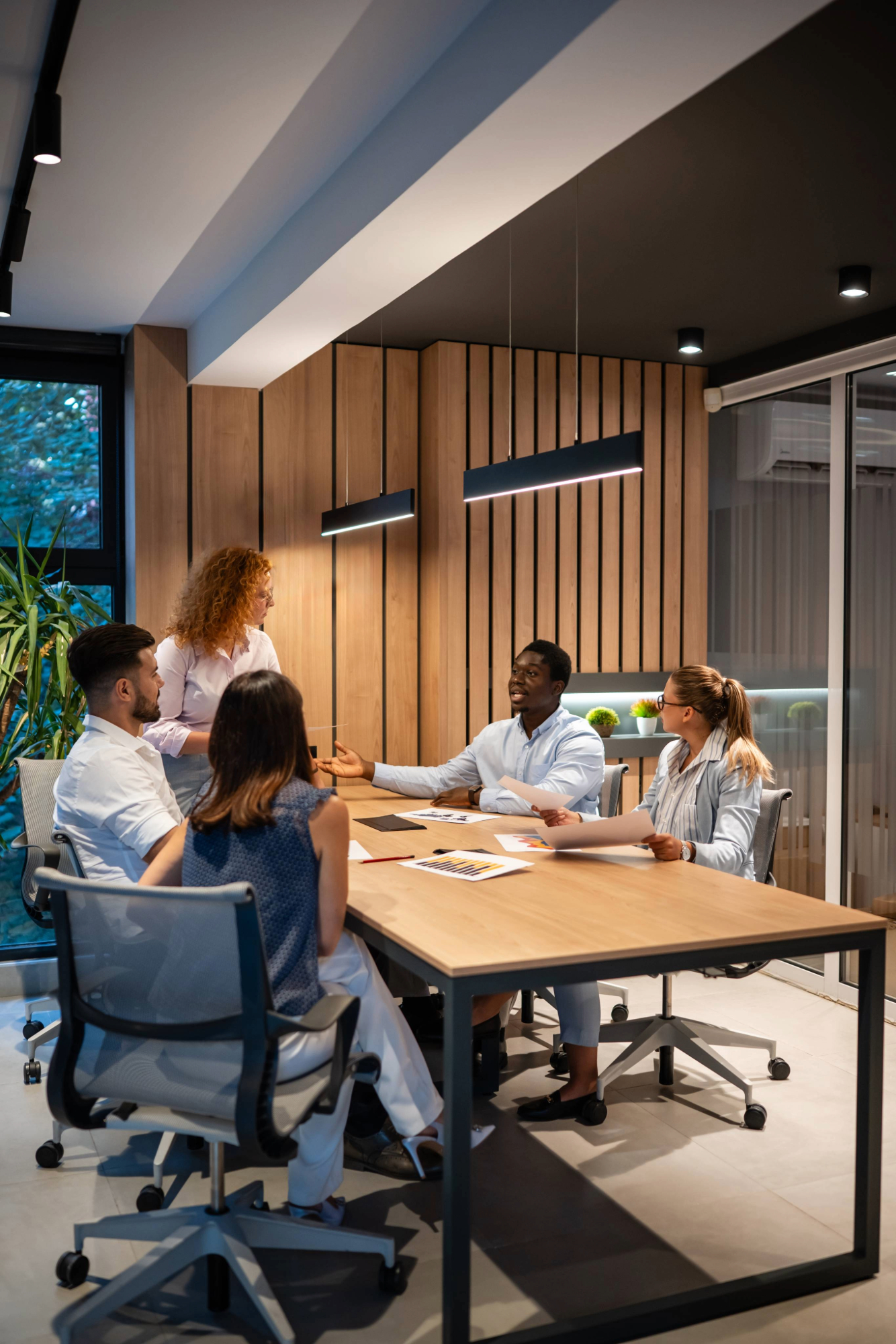 A group of 4 professionals sit around a table, 1 stands near the head of the table. They're animated in discussion.